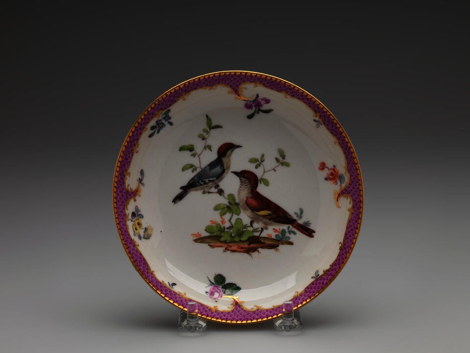A white saucer with gilded, pink, floral decorations, and two birds; one bird is blue with a red head positioned on a branch, the other is standing on the ground.