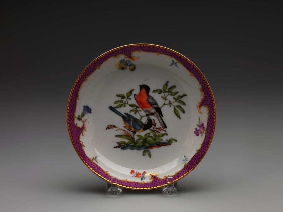 A white ceramic plate with a gilded and pink edge, floral patterns, with two birds surrounded by floral elements, positioned on branches, are blue, white, black, red, and yellow.