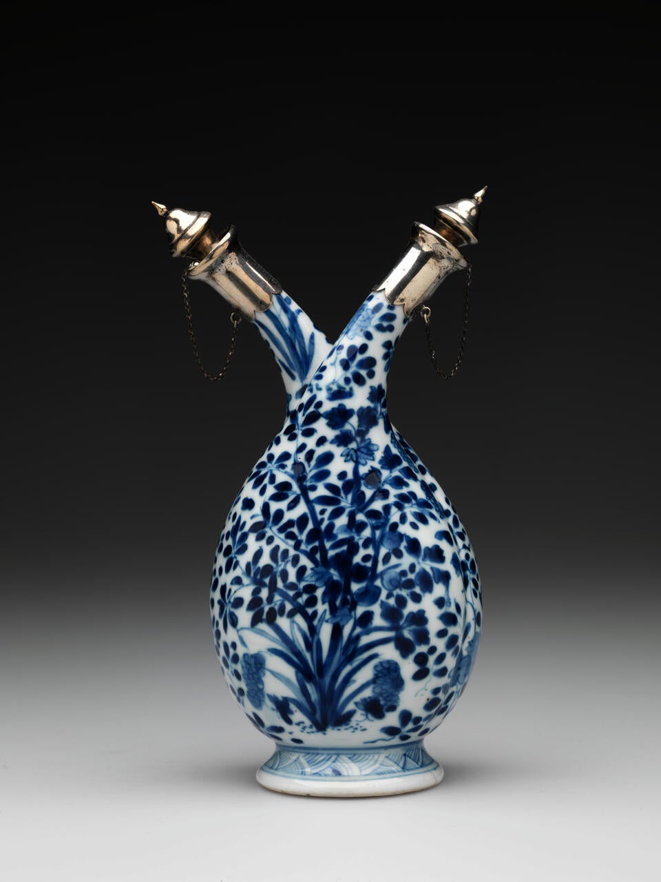 White and blue vessel with a flared foot, rounded body, and two necks in opposite directions. Necks are closed with silver caps attached to the vessel with delicate chains. 
