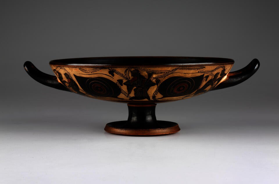 A wide, shallow, footed vessel with two handles on either side, curving upwards. The body is black and tan colored with an abstract design. 
