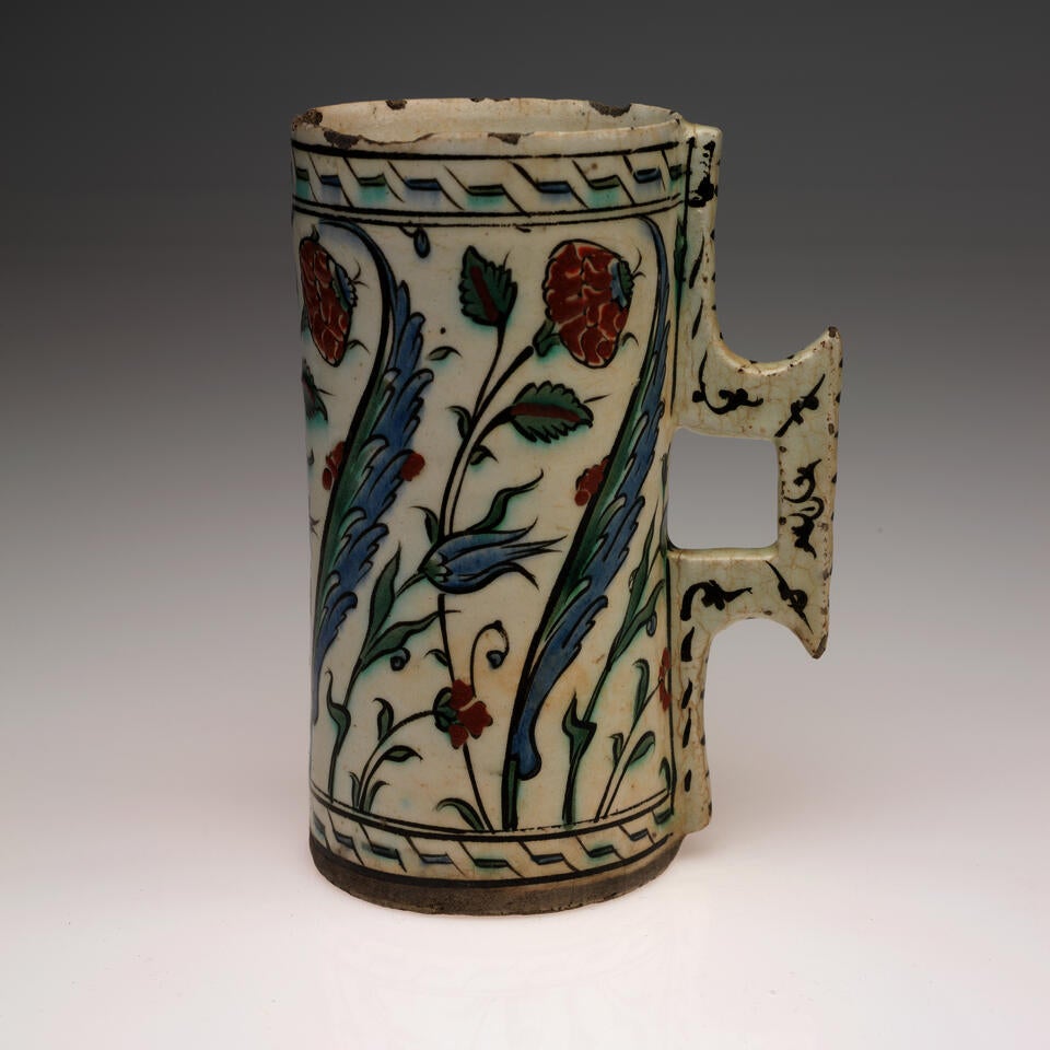 A rounded cream colored vessel with a square handle, decorated with geometric and flower designs in blue, green, and red. 
