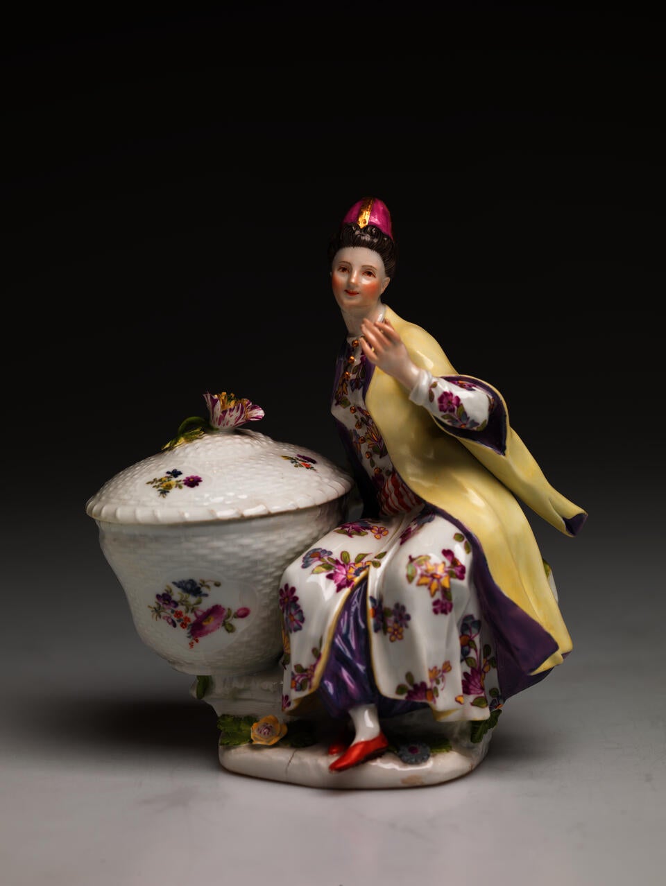 A sculptural figure sitting next to a large basket, the basket and figures clothing has floral decorations. The figure is wearing a hat, cream and purple clothing, and red shoes. 
