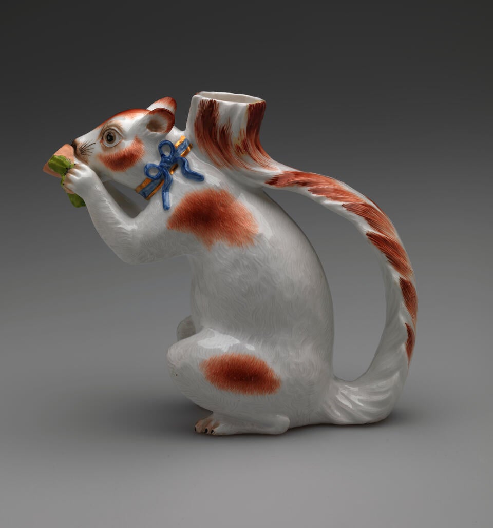 Vessel in the shape of a white squirrel with brown patches and a blue ribbon arounds its neck. It holds a brown nut with green leaves up to its face.