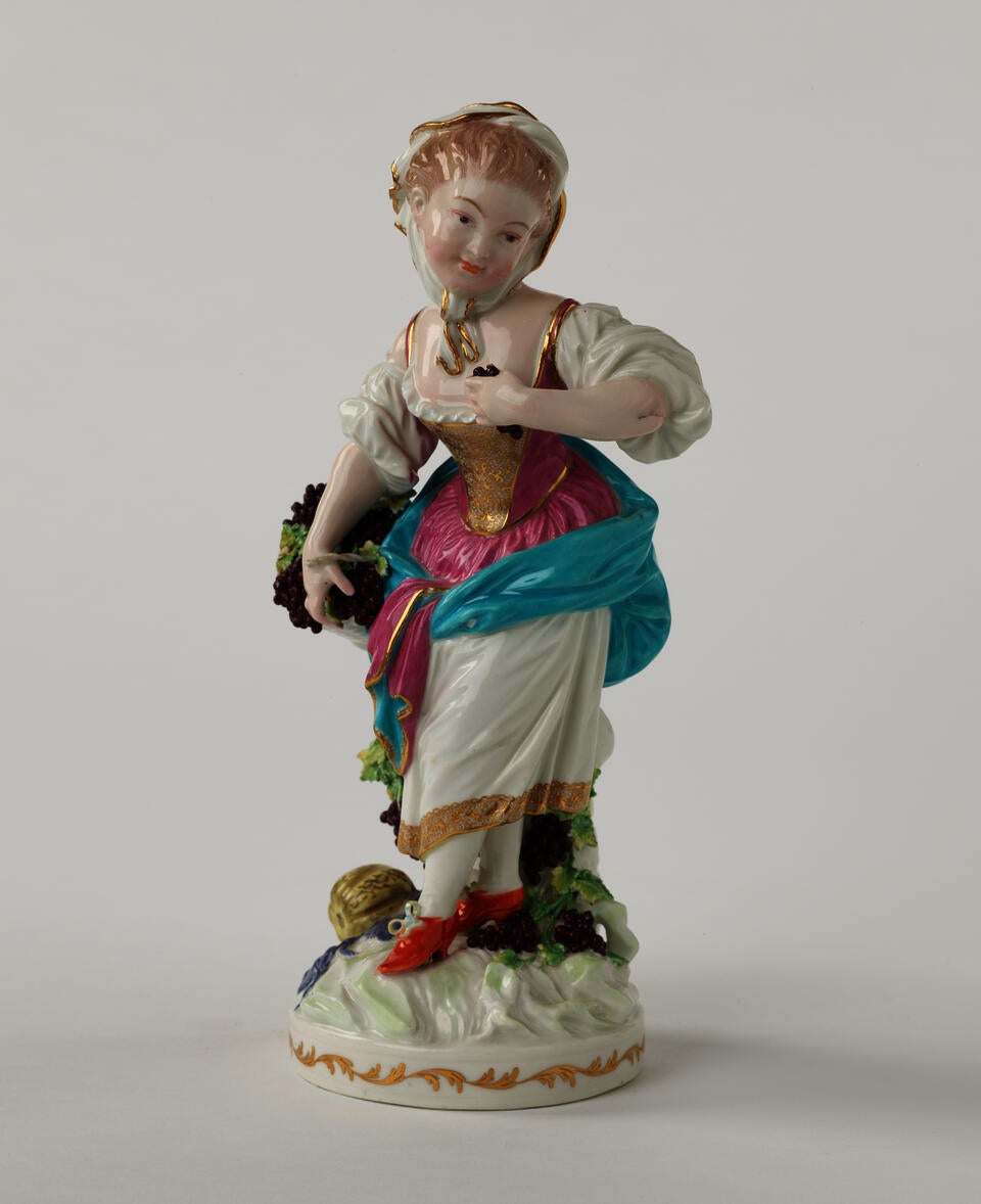 A sculpted porcelain light haired figure with grapes dressed in historical clothing that is gold, white, blue, pink, and red. 