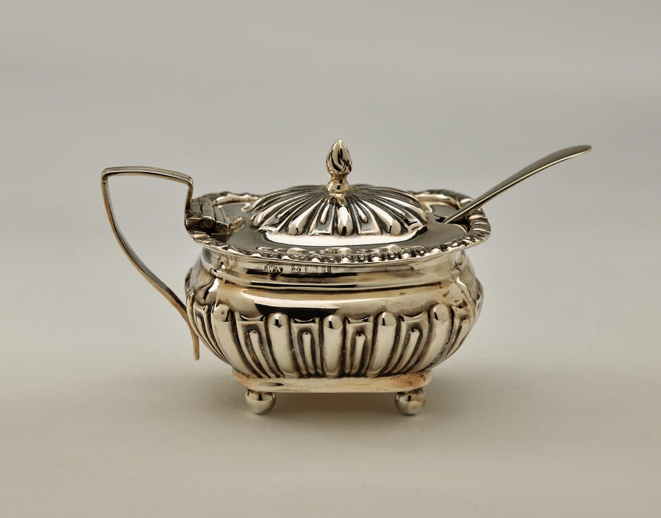  A silver, glass, and gilded mustard pot with a protruding spoon, the lid is hinged to the angular handle. The decorations are raised. 