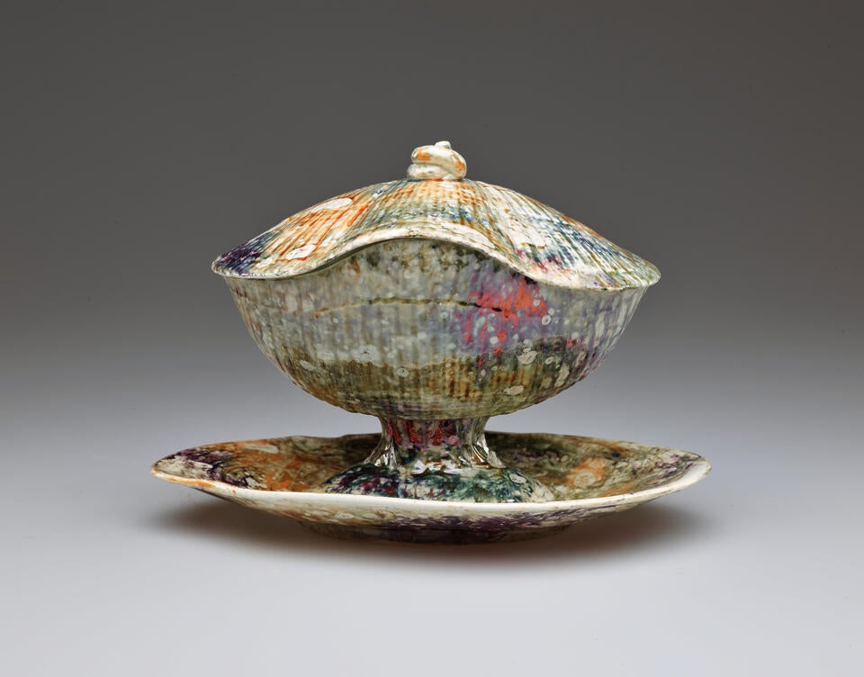 A lidded ceramic vessel on an organically shaped tray. A small, swirled finial is on the lid. The body of the vessel has ridges covered with a mottled, multi-colored glaze.