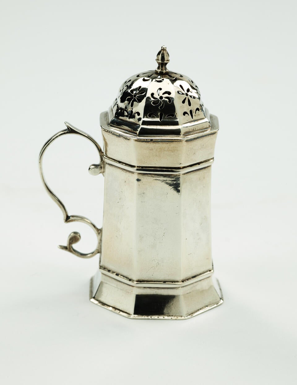 A silver caster that has an octagonal body, handle, and lid that has floral perforations.