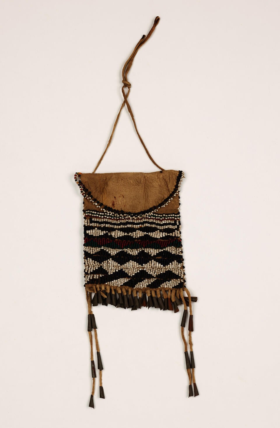 A leather beaded pouch with white, black, red, and green beaded designs. There is a handle as well as fringes with metal and leather decorations.