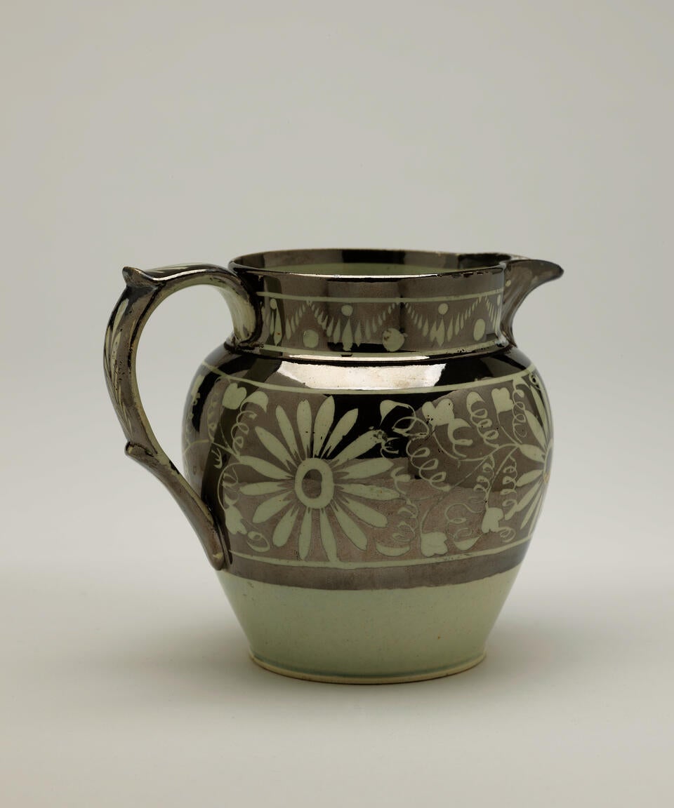 A silver ceramic jug with a decorative handle, there are decorative floral motifs on the body of the vessel.