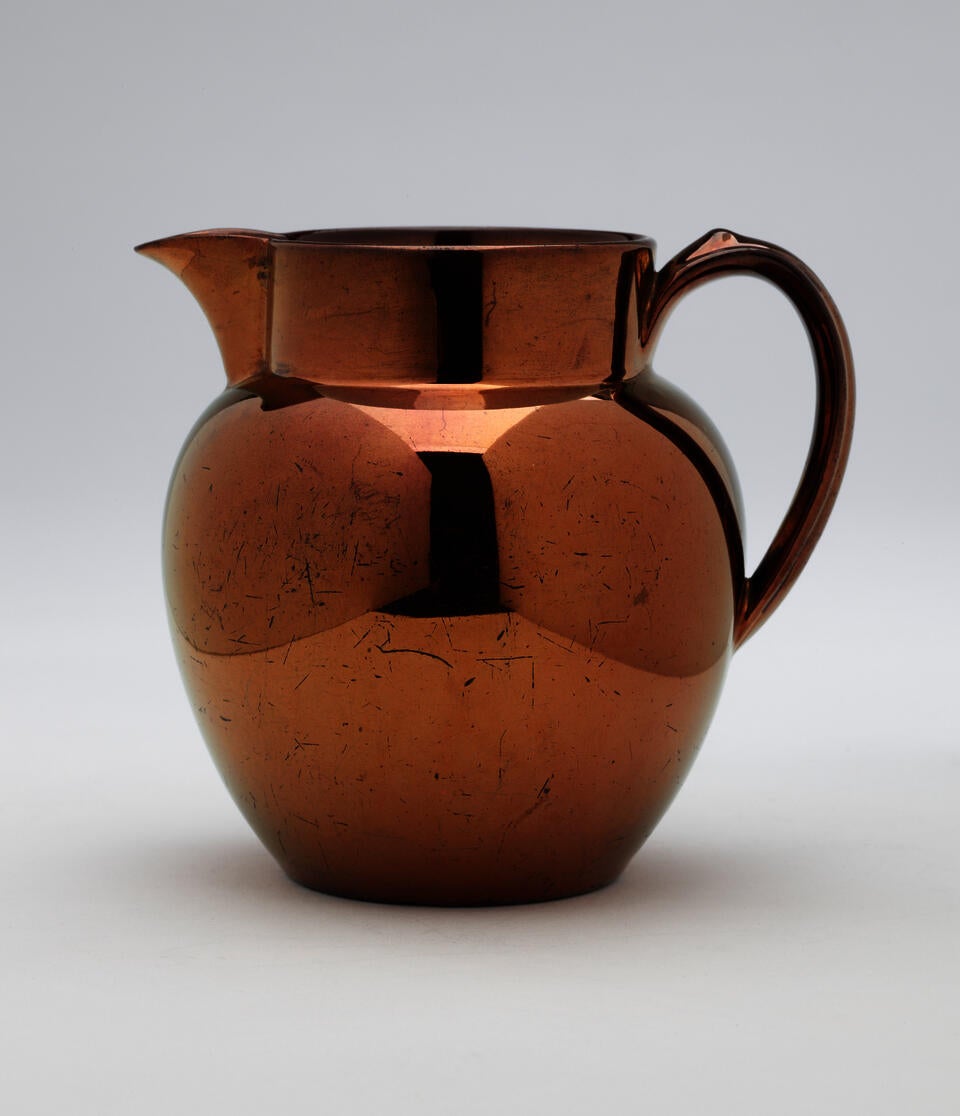 A ceramic jug with a small handle. The glaze is a brown metallic color.