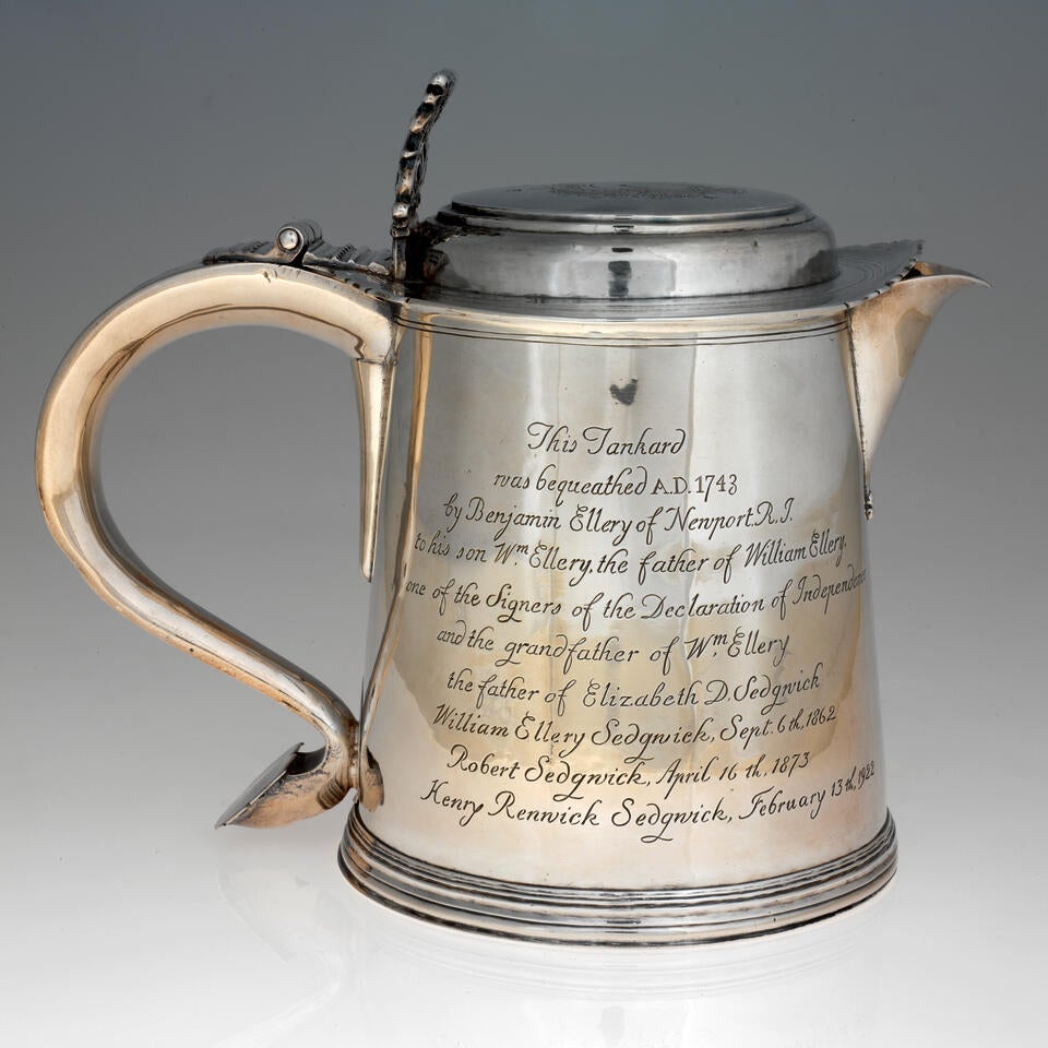 A rounded silver vessel with a curved handle, lid, and spout. Side of the vessel is covered with engraved writing. 