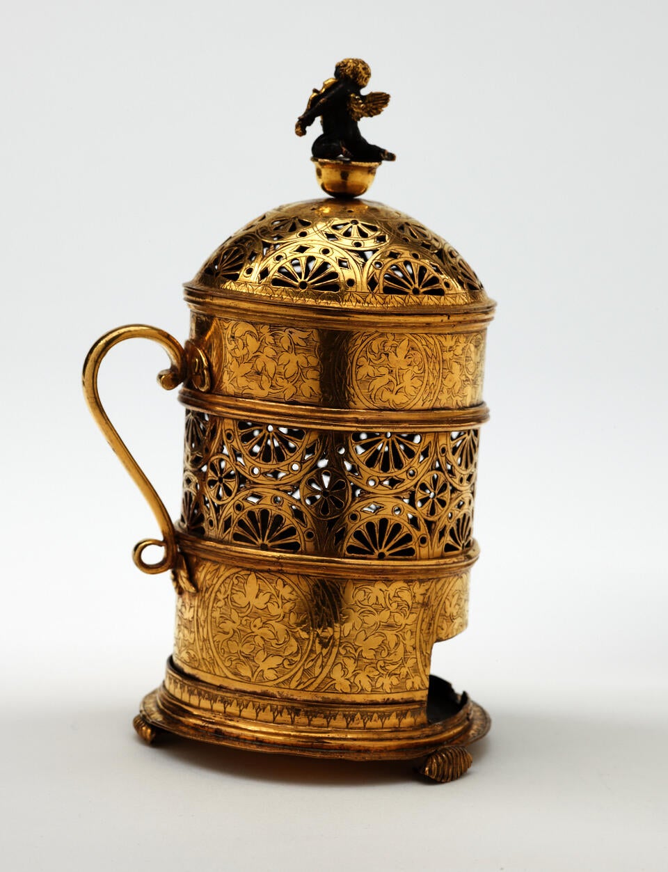 Gold colored pierced vessel with handle. Small dark figure sits on top.