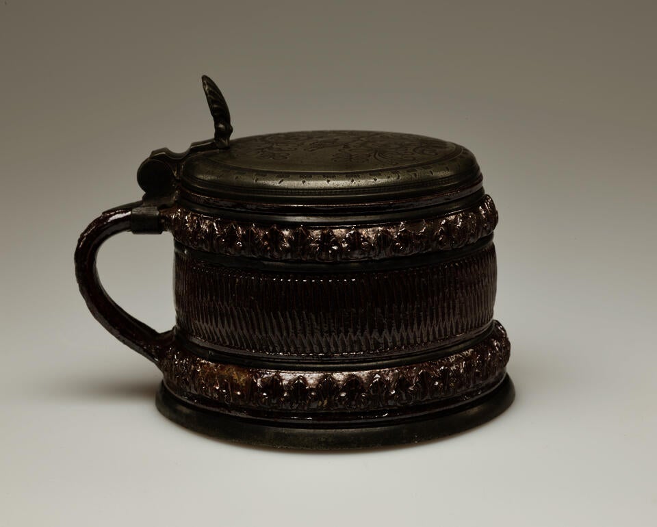 A short metallic glazed stoneware tankard with a delicate handle and hinged lid. There is abstract repeating decoration along bands on the entire vessel.