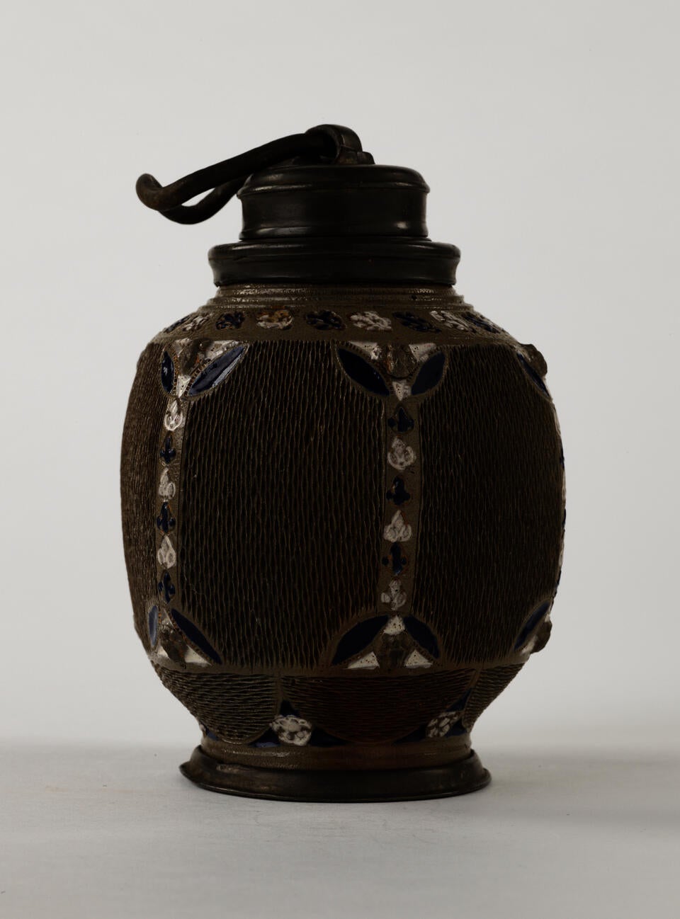A ceramic and salt glaze flask that is dark gray, blue, and white. The body is angular and has textured sections whereas the foot and top are rounded. 