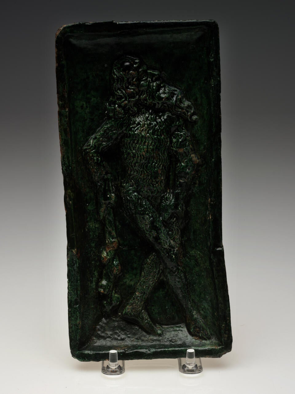 A green sculpted tile of a figure with long hair and beard, holding a stick-like object.