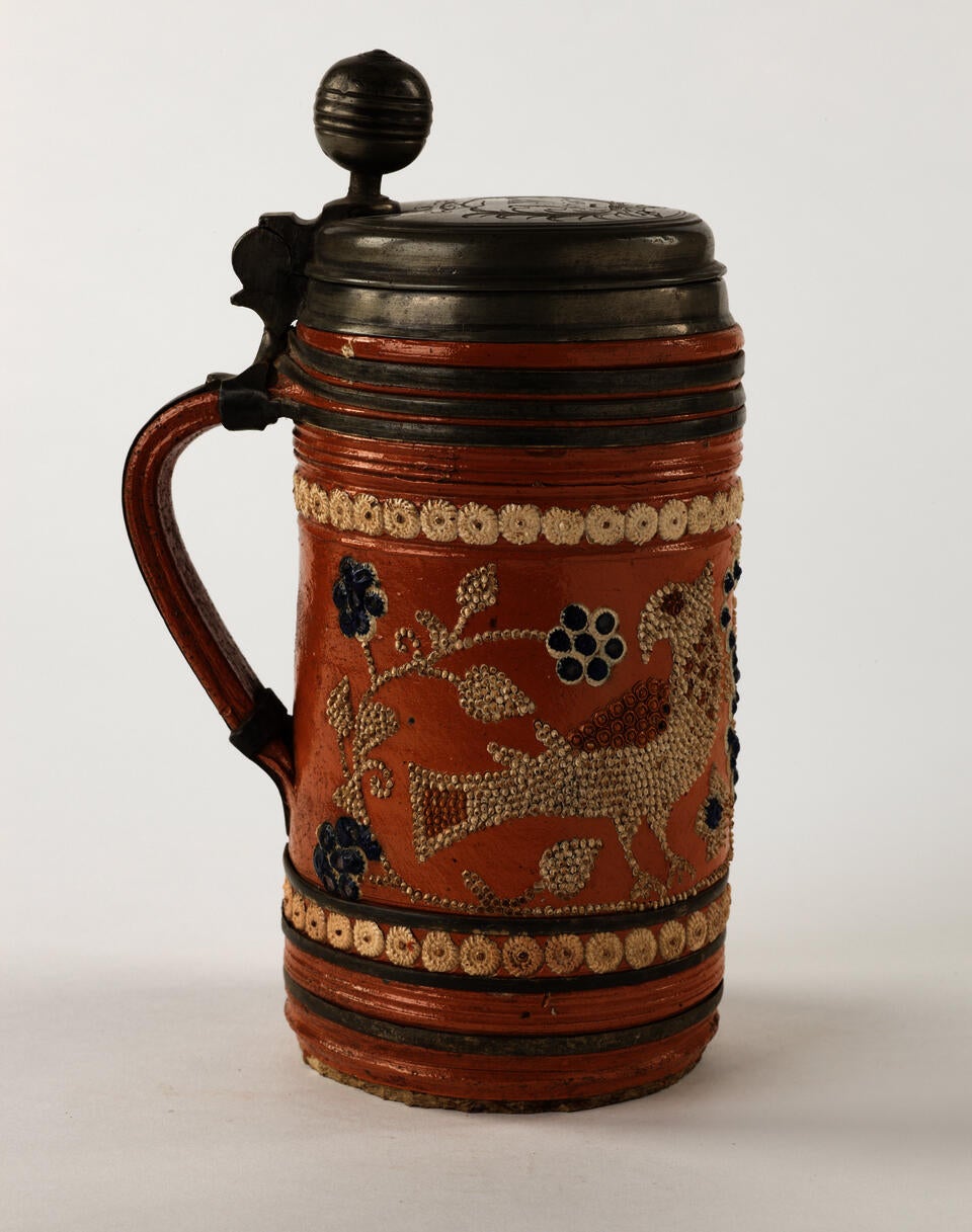 An orange earthenware stein with a metal lid that is hinged to the handle.  The white and blue decorations consist of dots and are primarily depicting floral imagery.