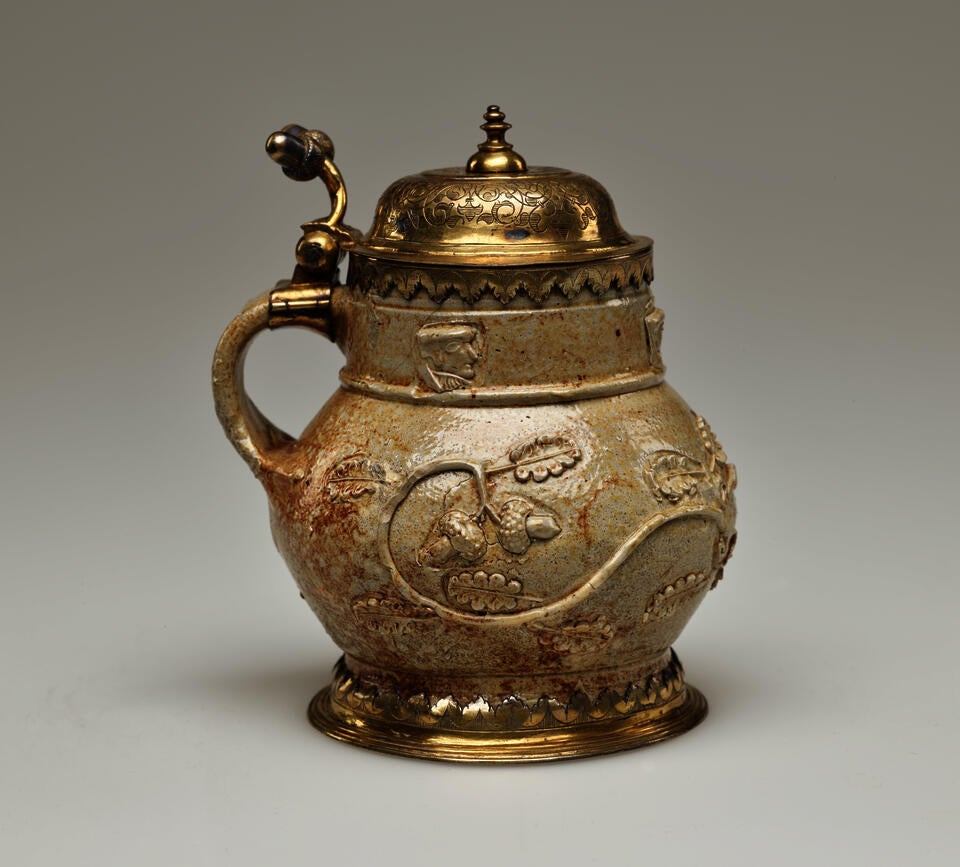 An unglazed stoneware mug with tan to copper colors and floral decoration. The foot goes into a gilded metal decoration, similarly to the handle where the hinged lid is attached.