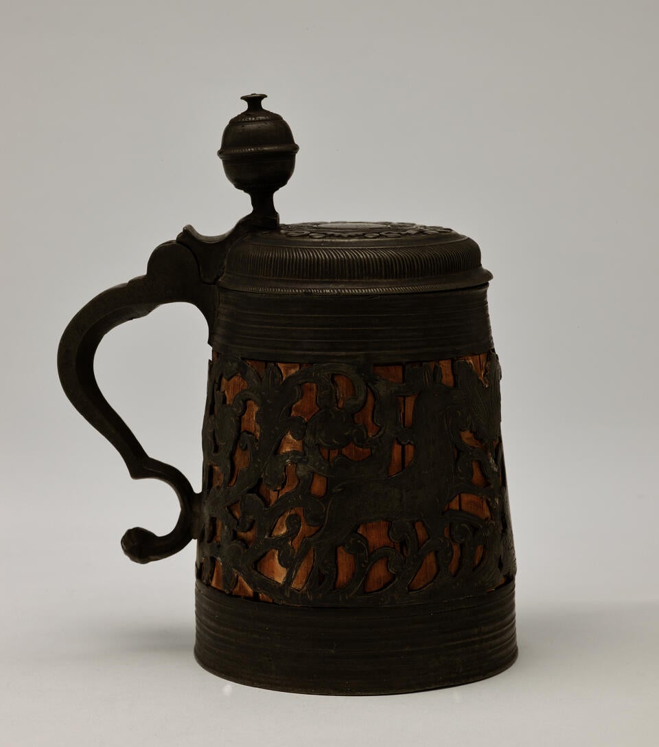 A wood and pewter mug that has a hinged lid and handle. The pewter is cut out and shows the wood on the interior. 