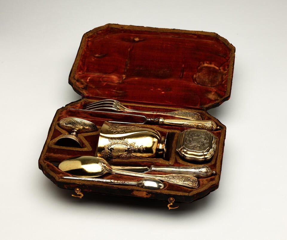 A traveling set with silverware. The traveling case is an organic colored fabric. There are multiple indents holding the silver, steel, and gilded cutlery and vessels.