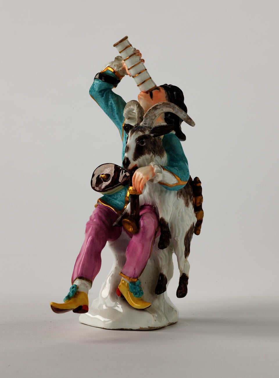 Sculpture of a light skinned man with a goat on his lap. He holds a dark brown instrument in one hand and drinks from a vessel in the other. 