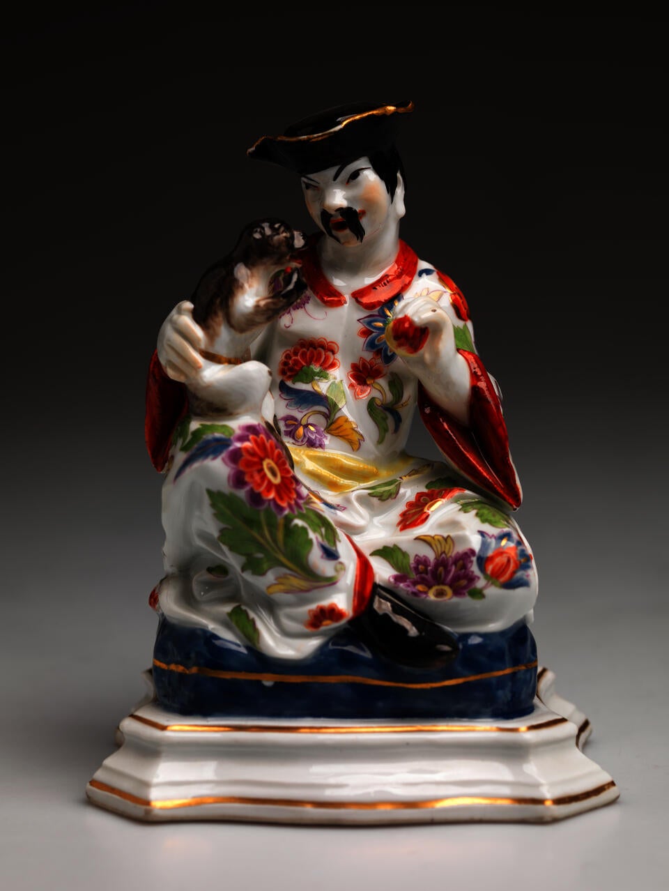 Sculpture of a caricatured Chinese figure wearing a floral garment and black hat. The figure holds a small animal on their lap.