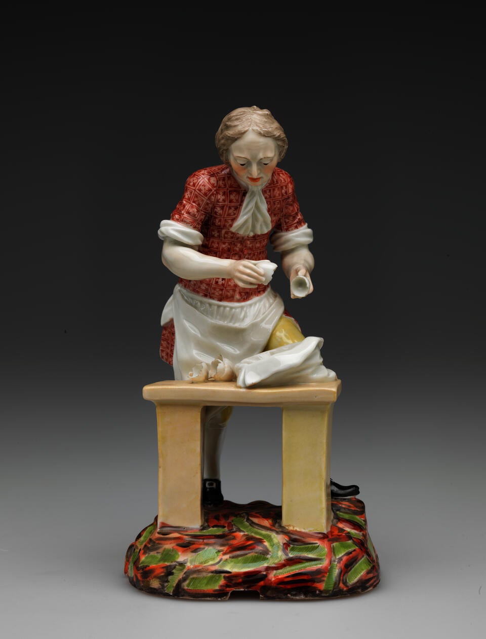 A sculpture of a figure in historical clothing and a table, a sack, and broken egg shells. The ground is brightly colored red, brown, and green. 
