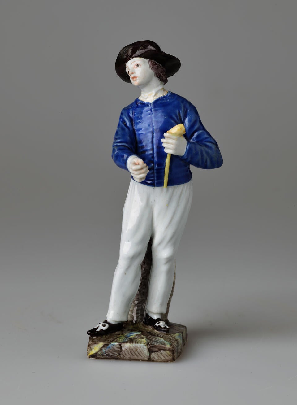 A sculpture of a figure in historical clothes, a blue shirt, white pants, black shoes and hat. The figure is also holding a pipe.