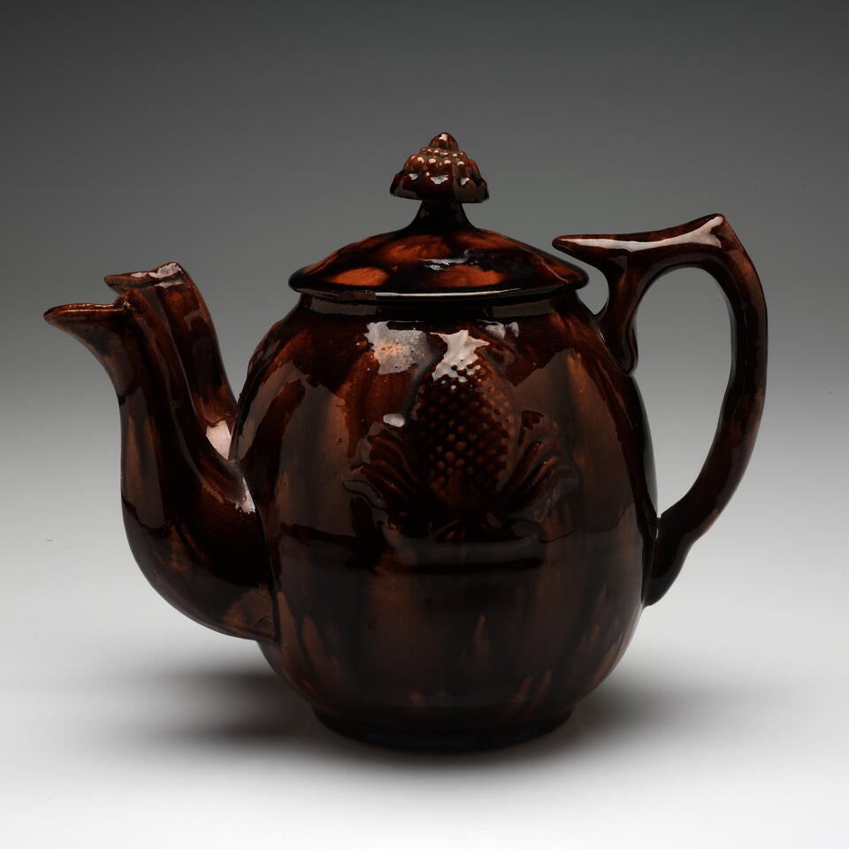 A mottled brown teapot with an image of a pineapple in the center. Two spouts are on the left side, and a rounded handle with a flat top faces right.