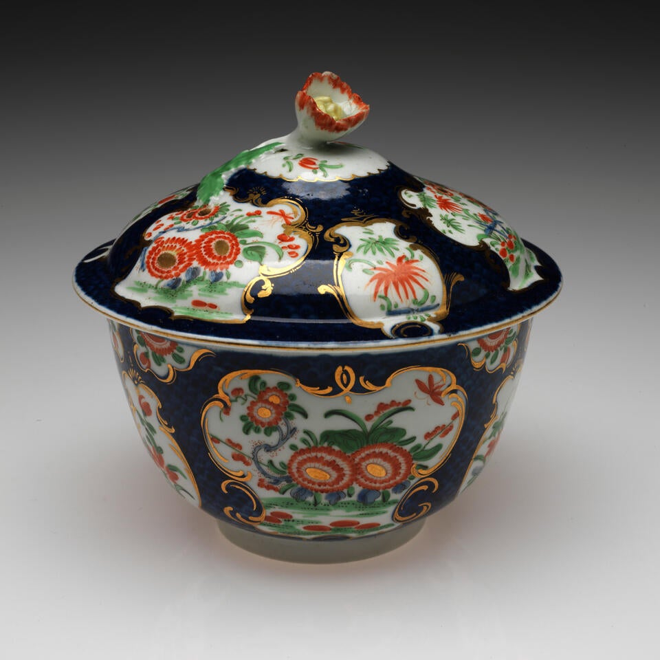 A lidded sugar bowl with floral decorations in gilded vignettes, surrounded by dark blue glaze, the finial on the lid is a sculptural red, white, and yellow flower. 