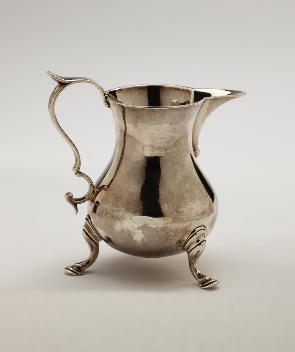 A silver creamer with a sculptural handle and three protruding feet. 