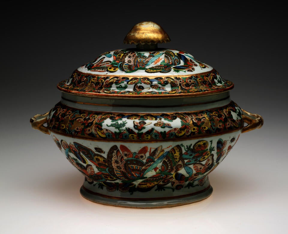 An oval lidded vessel with a large gold finial. The body of the vessel is decorated with multicolored butterflies on a white ground. 