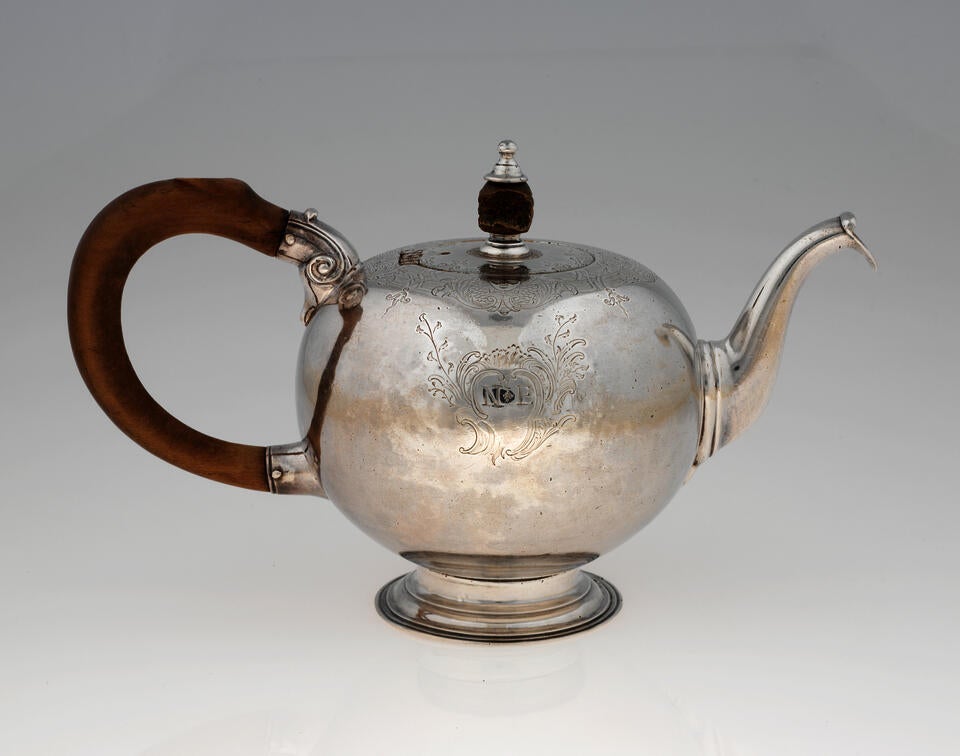 A bulbous silver teapot with a long curved spout, and foot, wooden finial and handle connected with decorative silver pieces. There are delicate engraved marks on the body and lid.