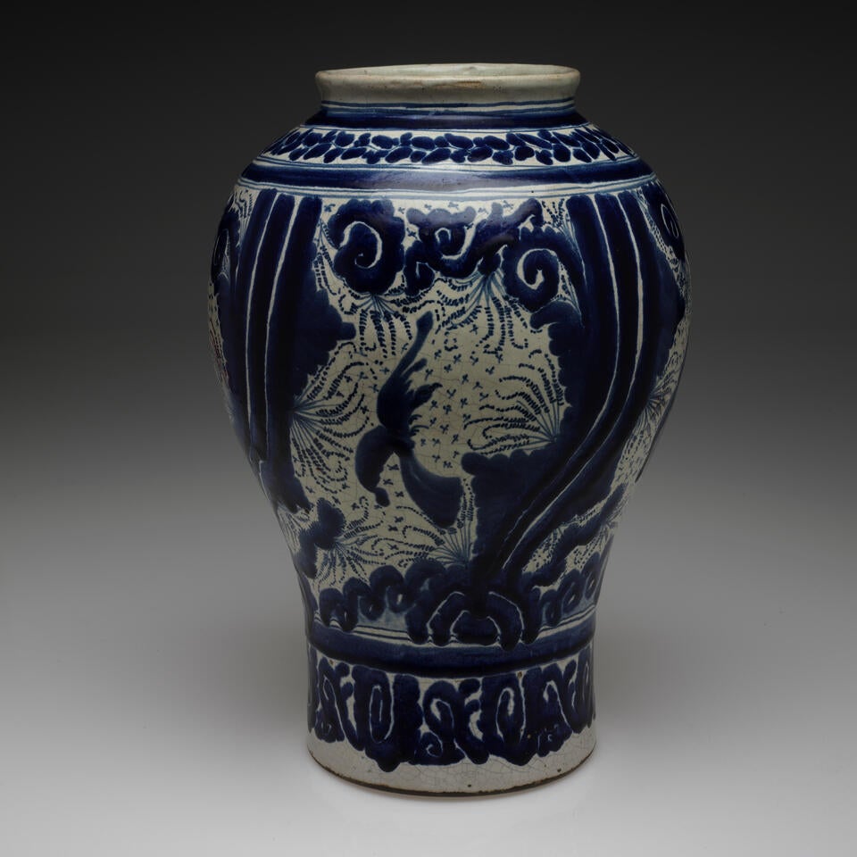 A vessel with a cylindrical bottom with a more bulbous top and a slightly flared out lip, it is glazed white and blue with abstract, purposeful and controlled decoration.