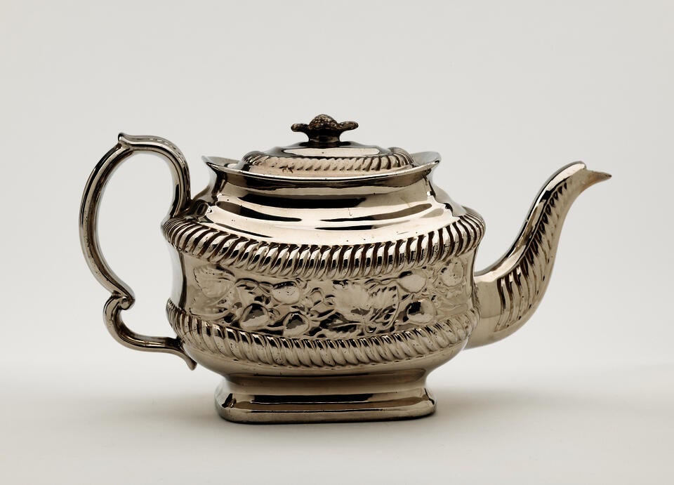 A silver luster teapot with a decorative handle, a rounded square body and spout with sculptural decorations.