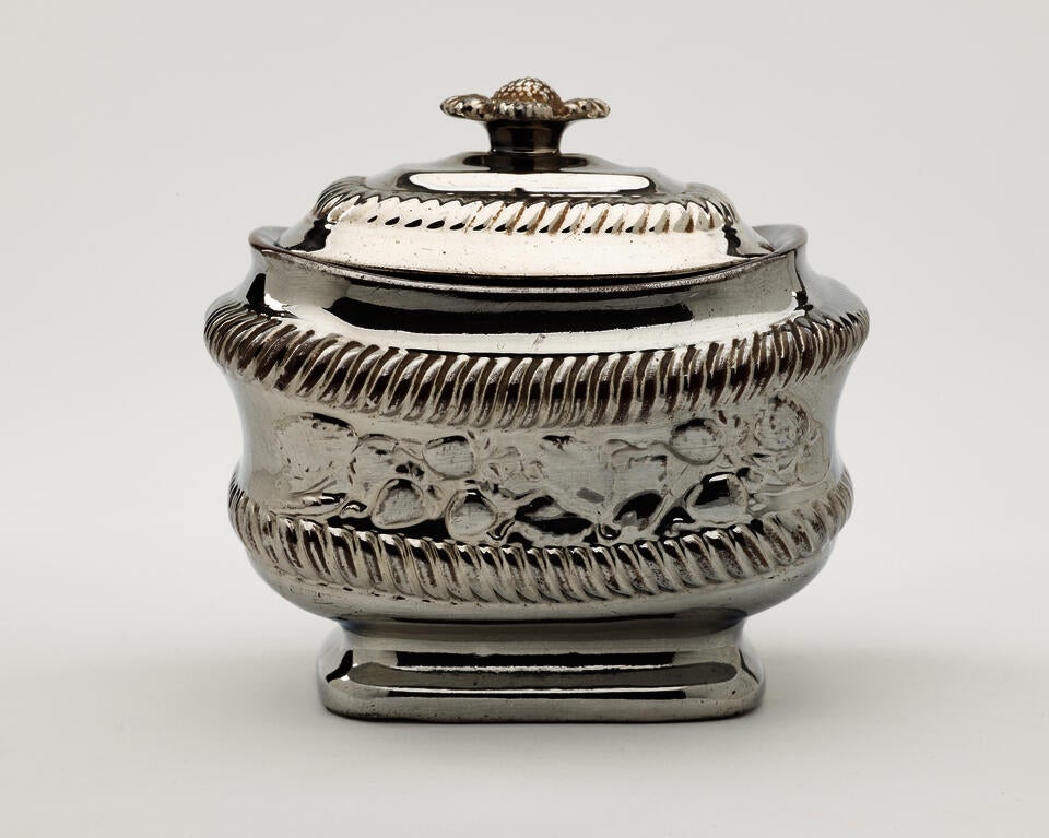 A silver luster sugar bowl with a decorative lid and a rounded square body with sculptural decorations.