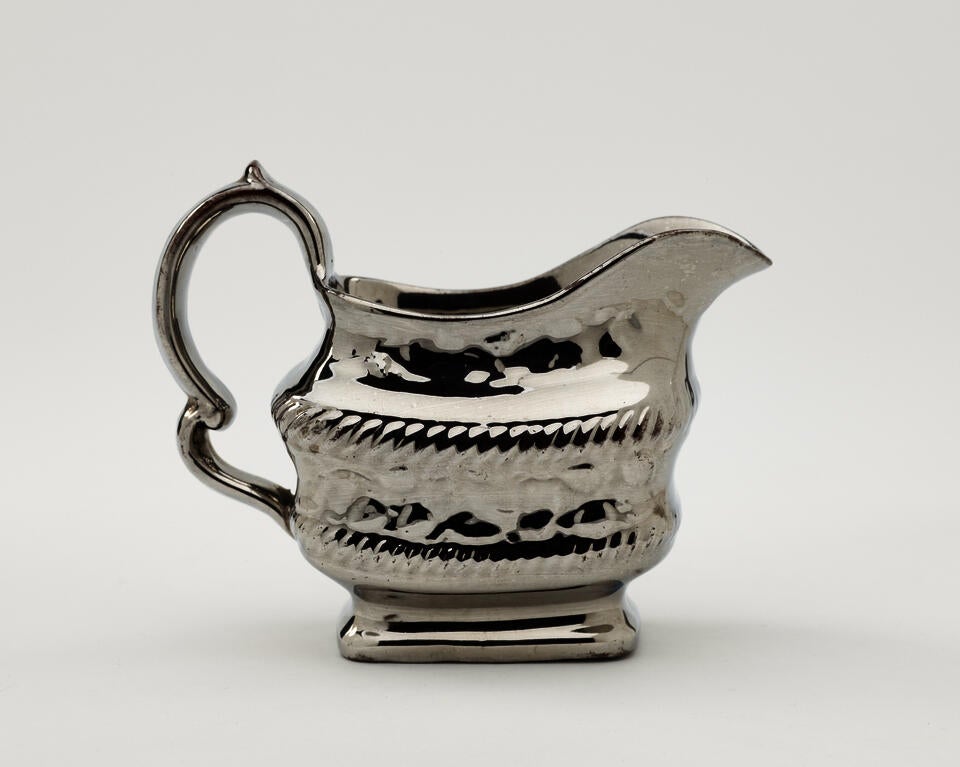 A silver luster creamer with a decorative handle, a rounded square body with sculptural decorations.