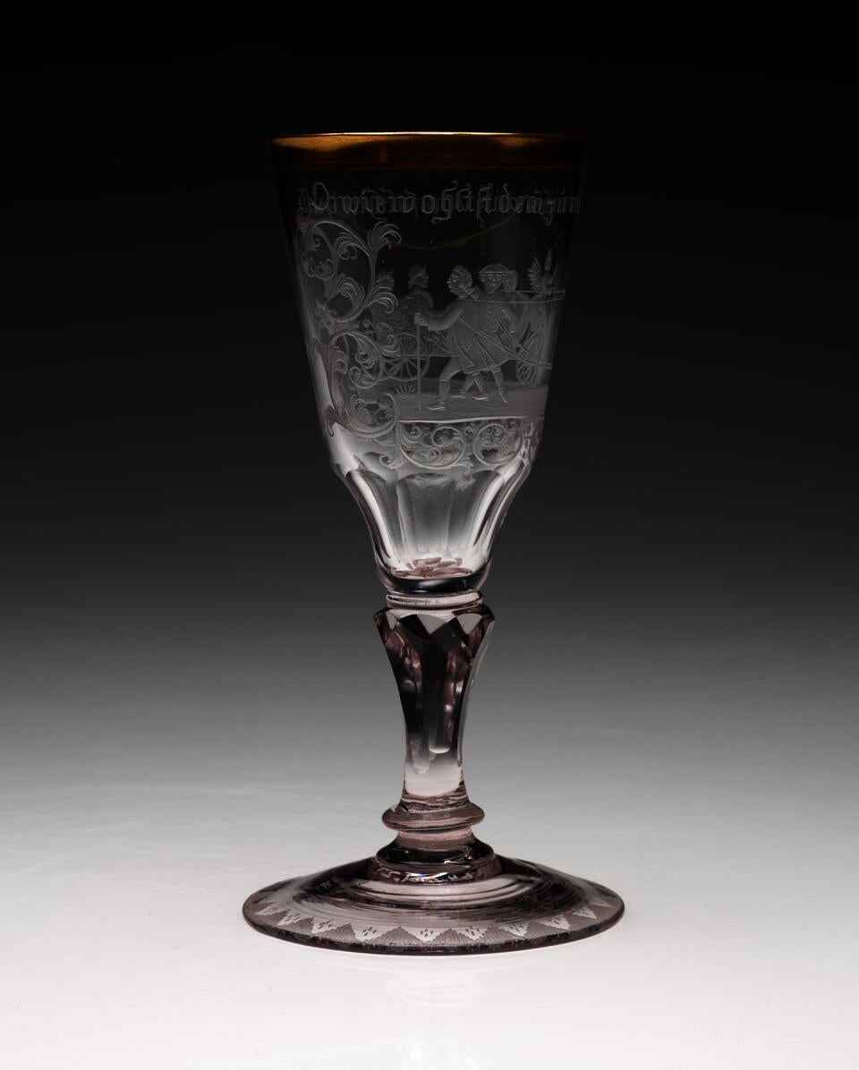 A clear stemmed glass with a rounded foot and thick gold rim. An image of figures walking in a group with words above them are etched in the sides. 