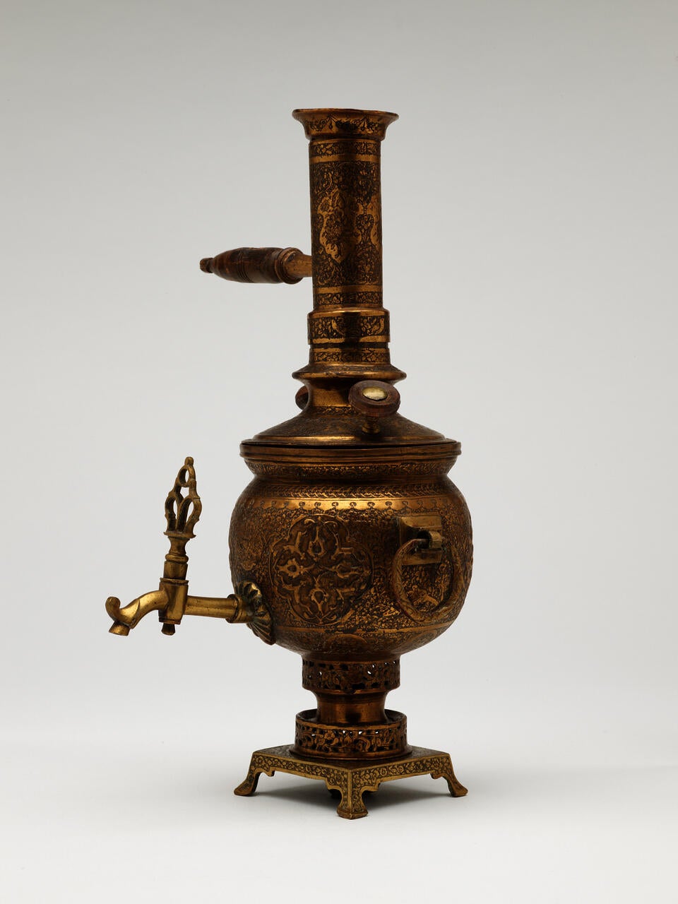 A brass and wood tea instrument that has a decorative foot, lid, various handles and rings, and spout. The entire vessel has highly decorative engravings. 