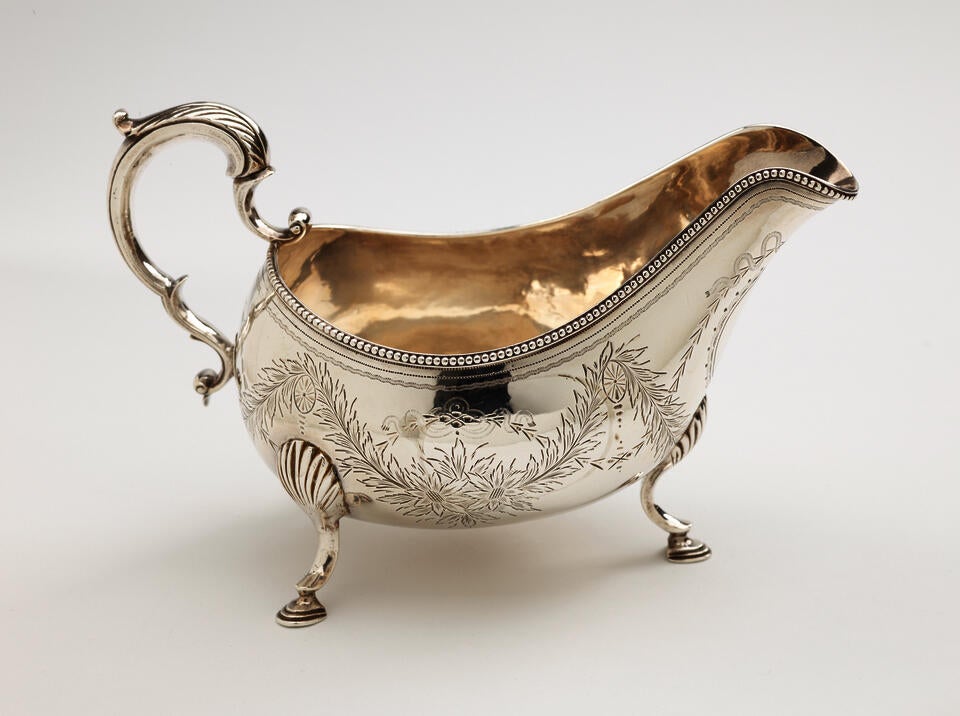 A silver sauce boat with a sculptural handle, high lip for pouring, and three protruding feet. There are floral engravings along the body of the vessel.
