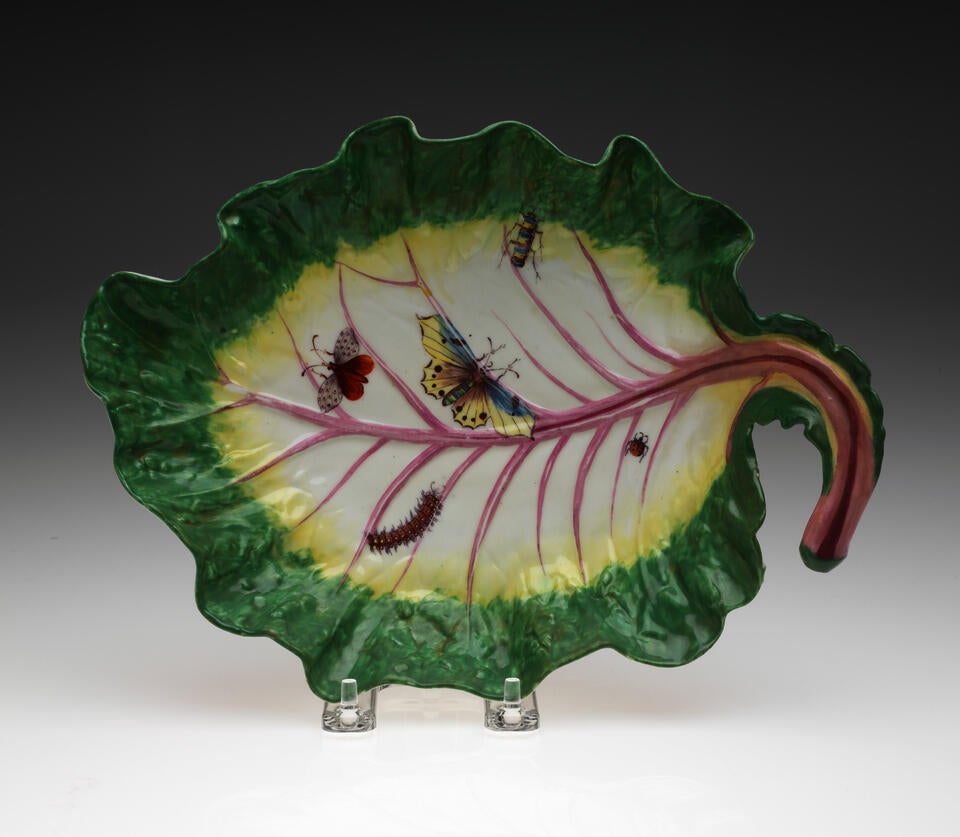 A leaf shaped porcelain dish, green on the edge to yellow then white, the stem is pink, with five insects depicted: a lady bug, two butterflies, a caterpillar, and beetle.