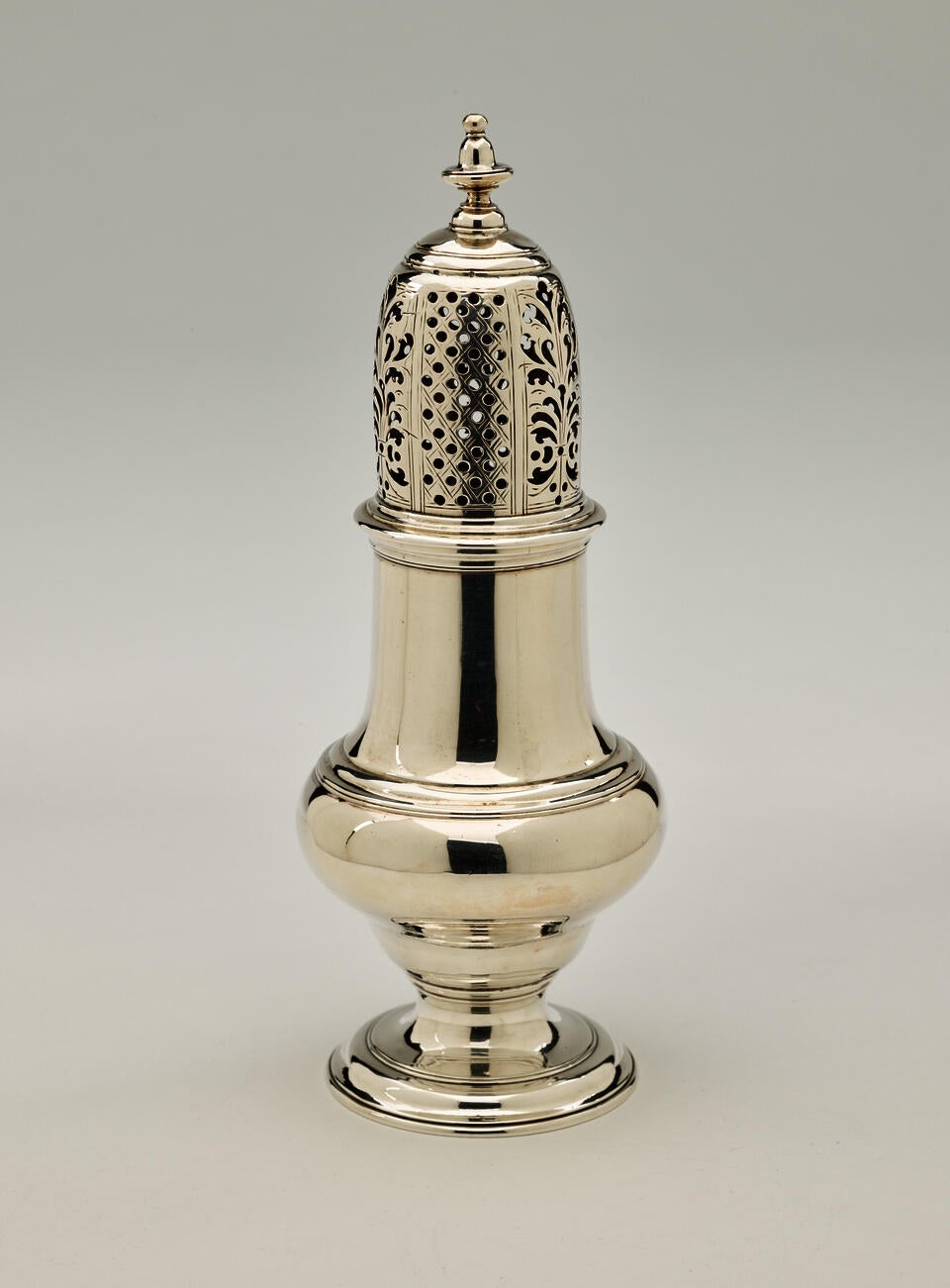 A silver muffineer with a foot, rounded body that goes to a cylindrical top half, and the lid is perforated with small holes and floral cutouts. 
