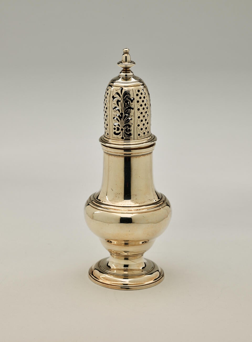 A silver muffineer with a foot, rounded body that goes to a cylindrical top half, and the lid is perforated with small holes and floral cutouts.