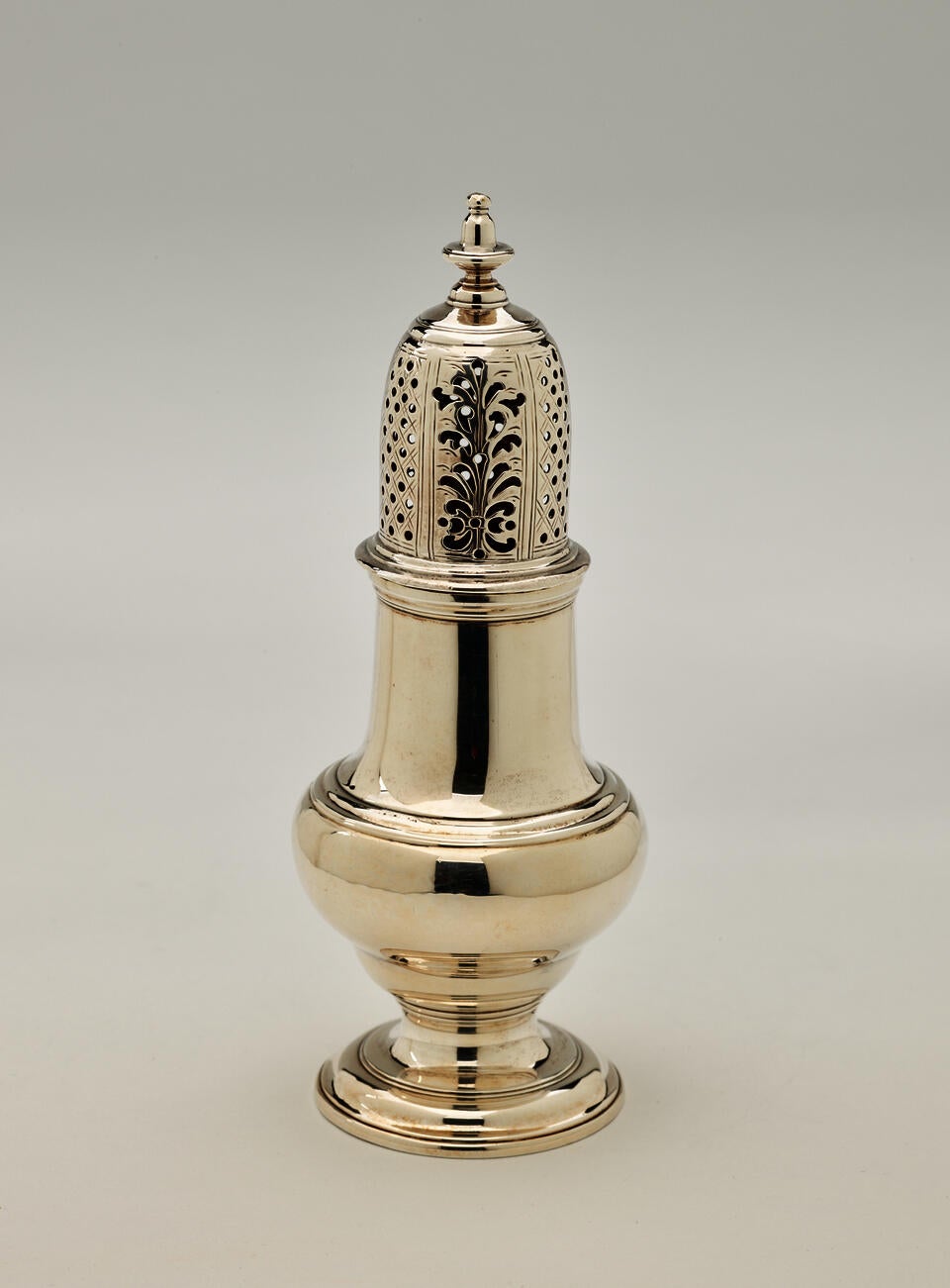 A silver muffineer with a foot, rounded body that goes to a cylindrical top half, and the lid is perforated with small holes and floral cutouts.