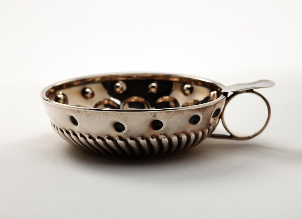 A silver wine taster with a simple circular handle, the body is very decorative.