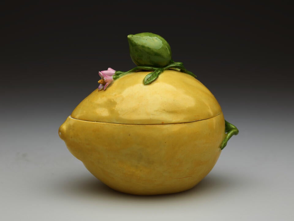 A porcelain lemon shaped box. It is light yellow and has sculpted floral elements such as a pink flower and green bud.  