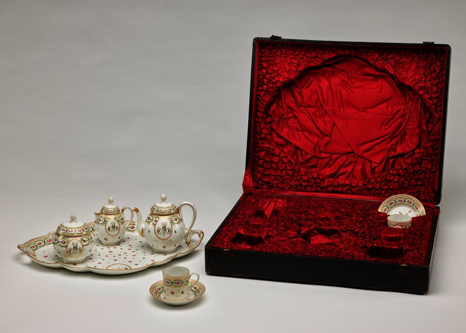 A white, pink, green, and gilded tea set with floral decorations and consists of four vessels and a tray. The traveling case with a black exterior and red fabric interior. 