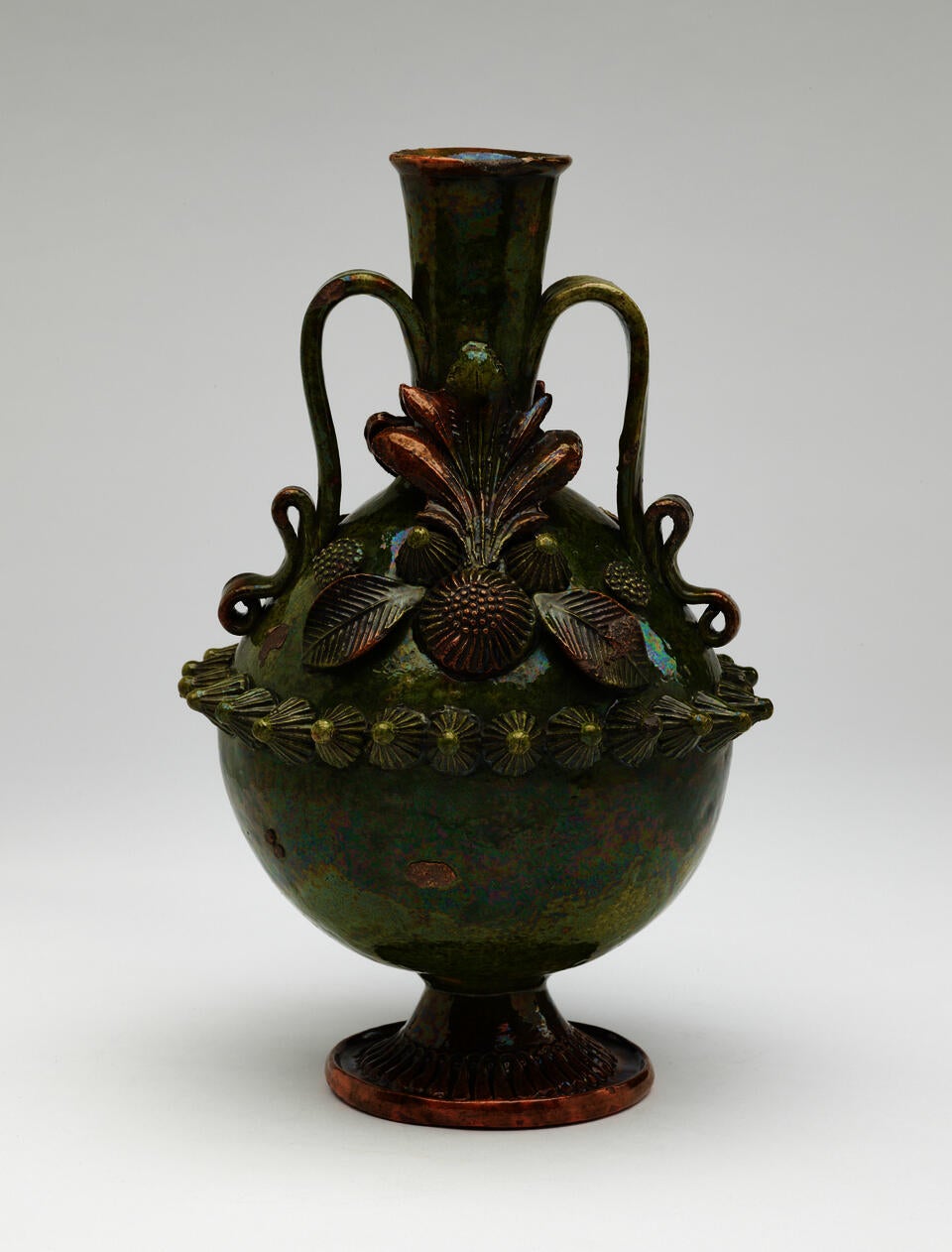 A dark green jug with sculpted decorations, symmetrical decorative handles, and a foot. 