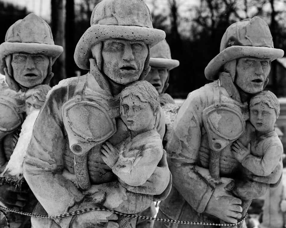 A group of identical white statues of firefighters carrying a child. A rope at the bottom of the frame binds them together.