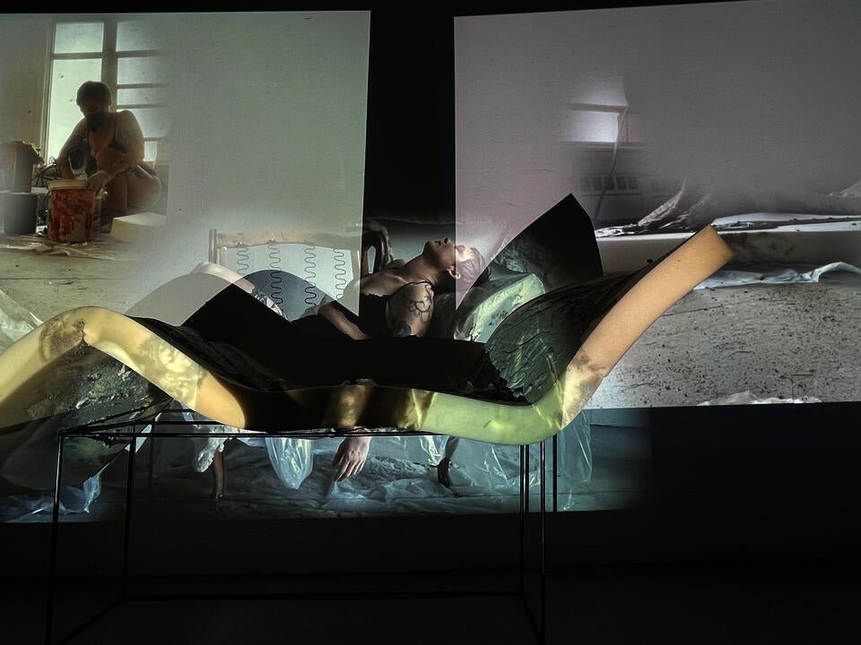 A Triptych of Projections showing the process of pouring concrete on foam, draping the foam across the arms of a chair and then lying over it. These three projections are over a wall and the hardened concrete foam piece. 
