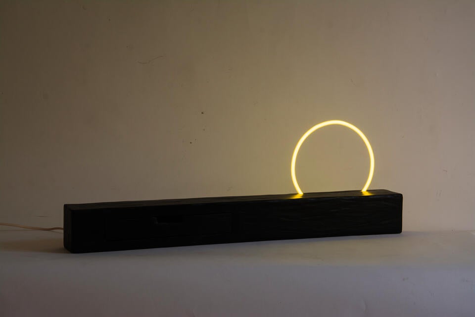 A long rectangular wooden form with an organic carved texture. A perfectly circular yellow neon protrudes from the top of the shelf. 